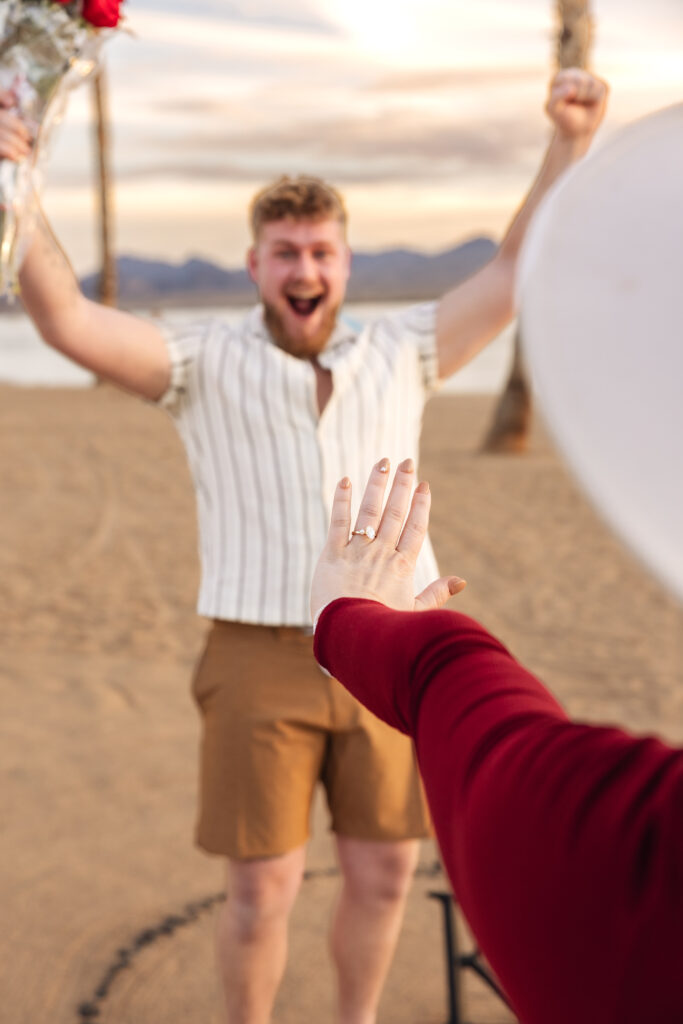 Woman showing off engagement ring on beach in Lake Havasu with fiancé cheering in the background