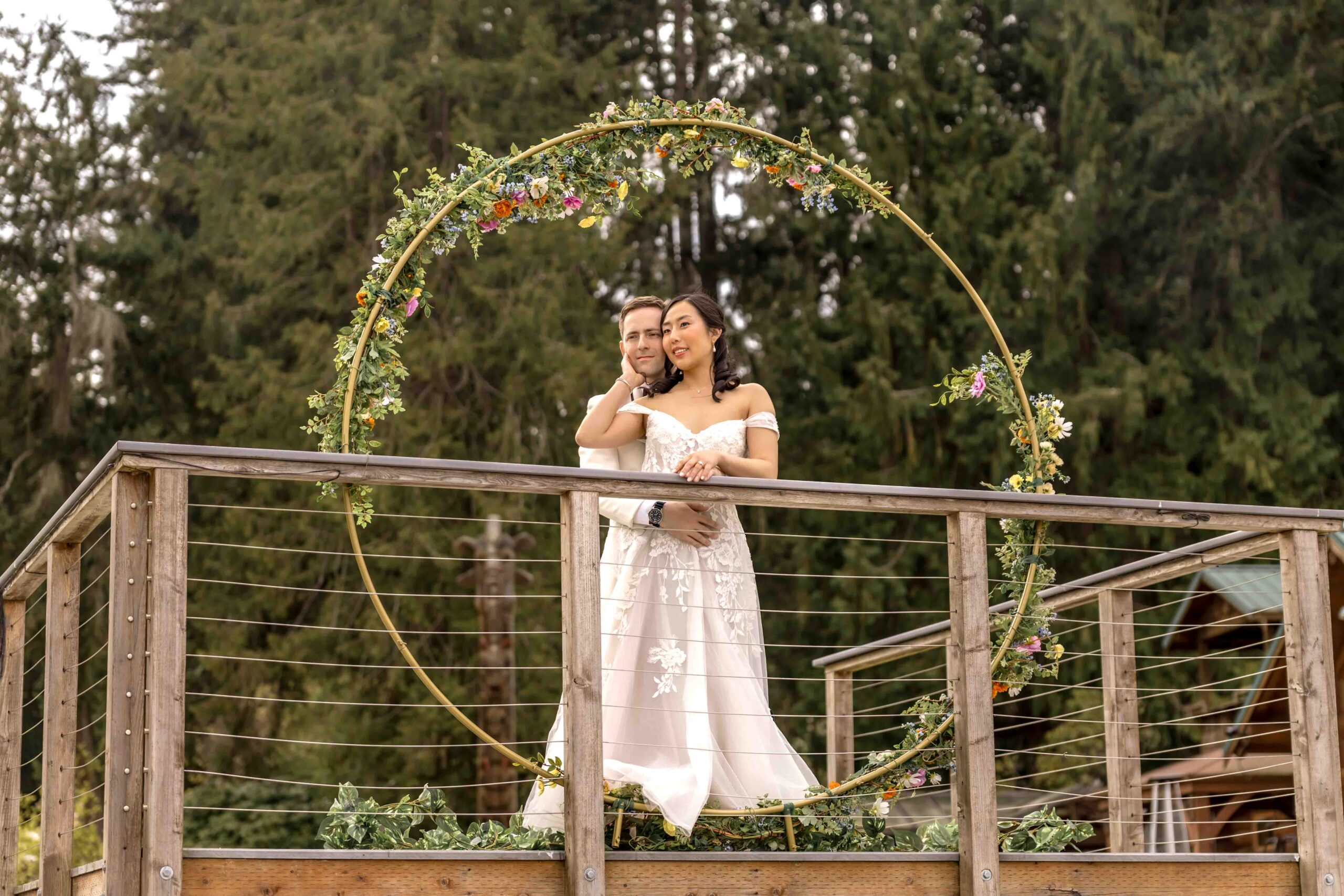 Bride and groom embrace under a floral circle arch on a wooden deck at Kiana Lodge in Seattle