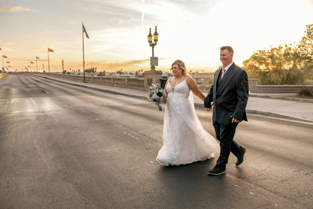 Bride and groom walking hand-in-hand across the London Bridge at sunset during their Lake Havasu wedding, captured by Elona Shea Photography.