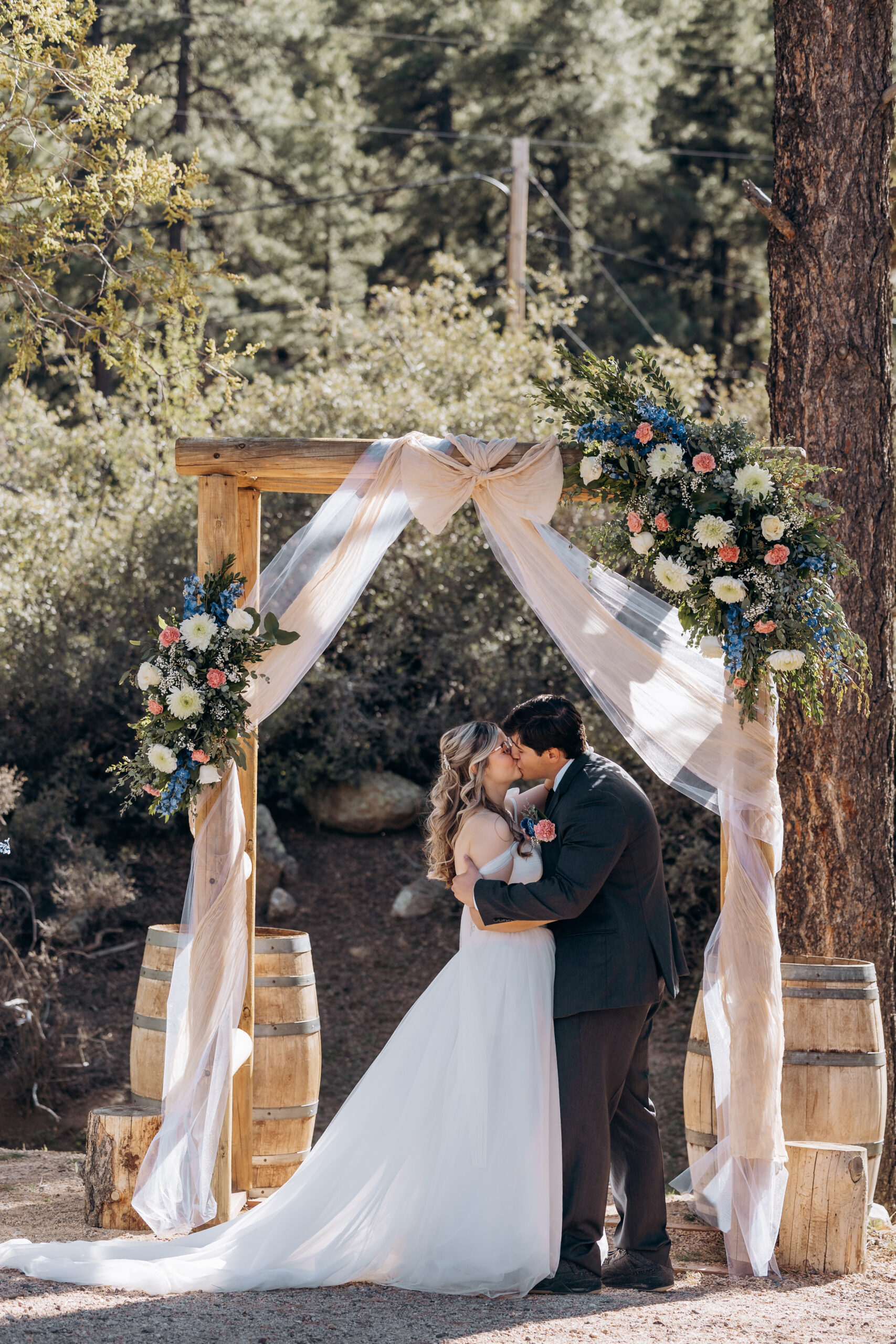 Bride and groom kissing under a wooden wedding arch with soft draping and floral arrangements during an outdoor ceremony
